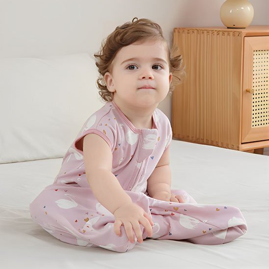 Bébé assis près d’un meuble en bois, portant une gigoteuse rose.