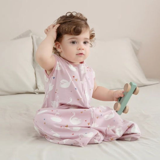 Bébé assis sur un lit, portant une gigoteuse rose et tenant un petit jouet vert