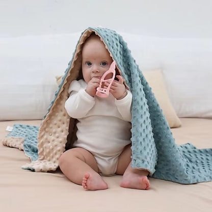 Petit bébé assis sur un canapé, enveloppé dans une couverture double-face beigne beige et bleu, tenant un petit jouet rose.
