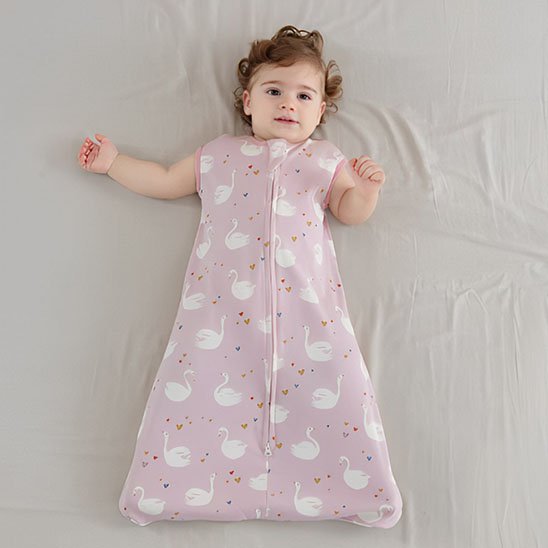 Bébé calme allongé sur un lit, vêtu d’une gigoteuse rose à motifs de cygnes blancs