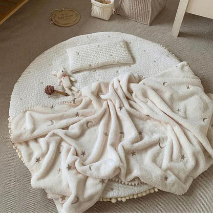 Couverture bebe crème avec étoiles et lunes brodées posée dans un espace de jeu pour bébé